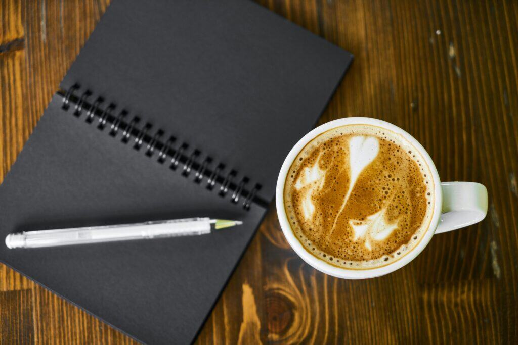 Open black notebook with a pen and a cup of coffee on a wooden desk, symbolizing journaling and mindfulness for consultants.