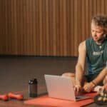 Consultant working on laptop while sitting on gym mat with exercise equipment nearby, demonstrating integration of health and fitness into business routine for better performance and productivity