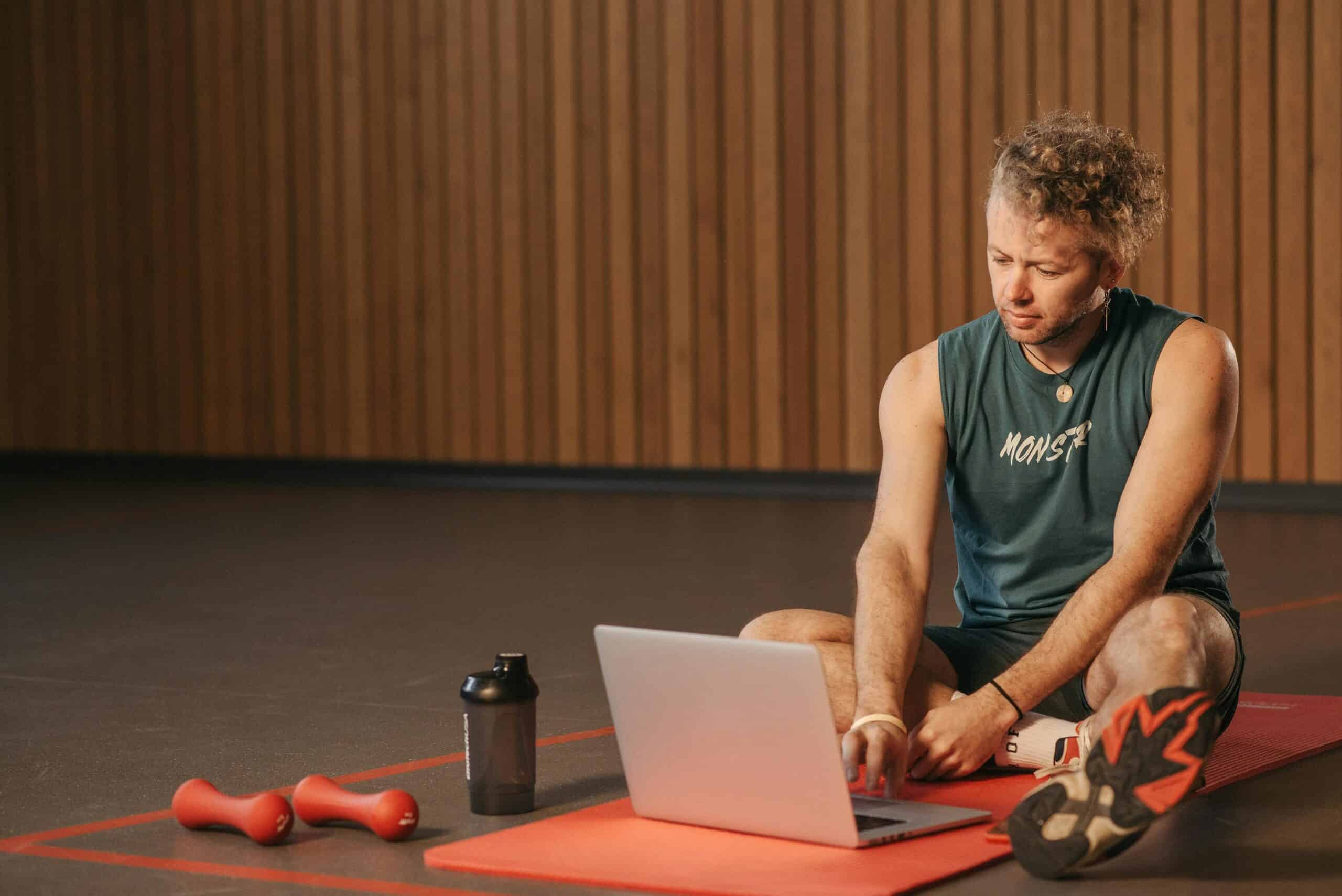 Consultant working on laptop while sitting on gym mat with exercise equipment nearby, demonstrating integration of health and fitness into business routine for better performance and productivity