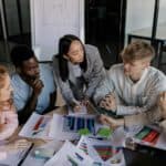 A small team in a modern office leans over a table covered in charts, graphs, and notes as they focus on planning and deciding their next business actions.