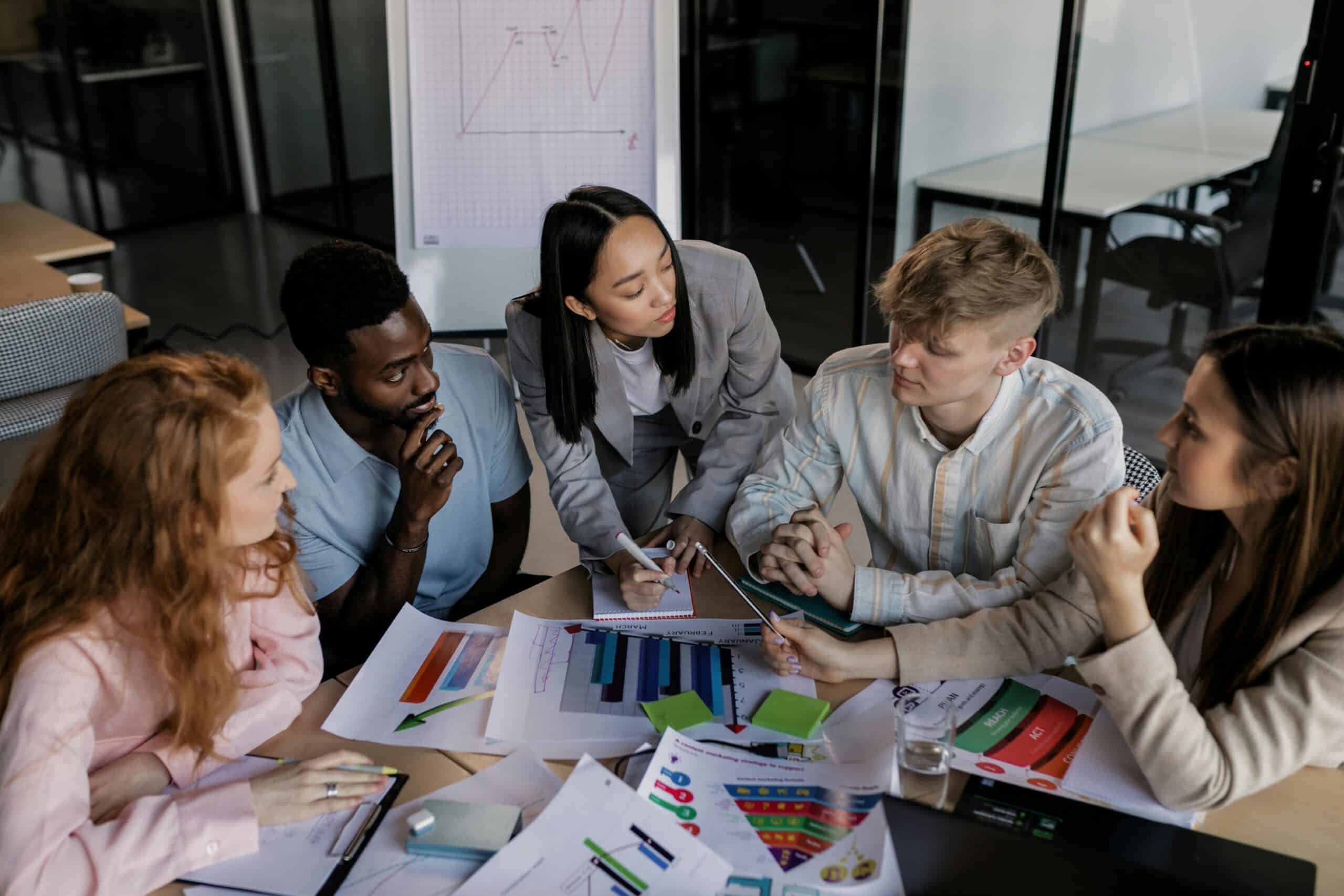 A small team in a modern office leans over a table covered in charts, graphs, and notes as they focus on planning and deciding their next business actions.