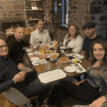 How to Choose the Right Consulting Firm Organizational Structure Group of six consulting colleagues smiling around a rustic wooden table at a restaurant, sharing a dinner of shared plates and wine in a warmly lit stone-walled dining room after a mastermind event.
