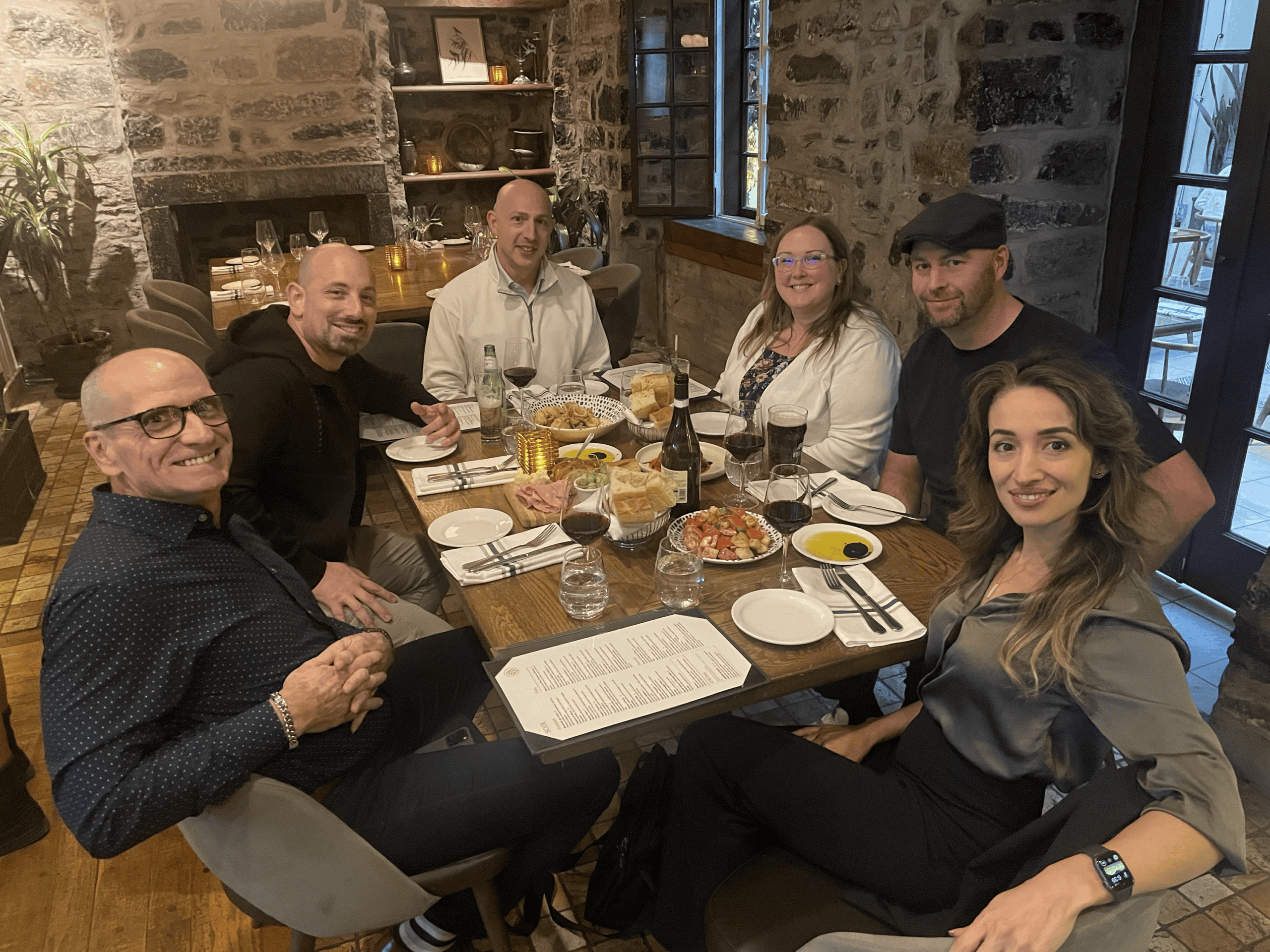 Group of six consulting colleagues smiling around a rustic wooden table at a restaurant, sharing a dinner of shared plates and wine in a warmly lit stone-walled dining room after a mastermind event.