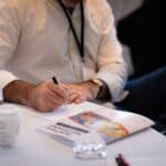 The Defaults Quietly Running Your Consulting Business Consultant in a white shirt and event lanyard writing notes by hand at a Consulting Success Montreal Mastermind, with a coffee, a glass of water, and an event guide on the table.