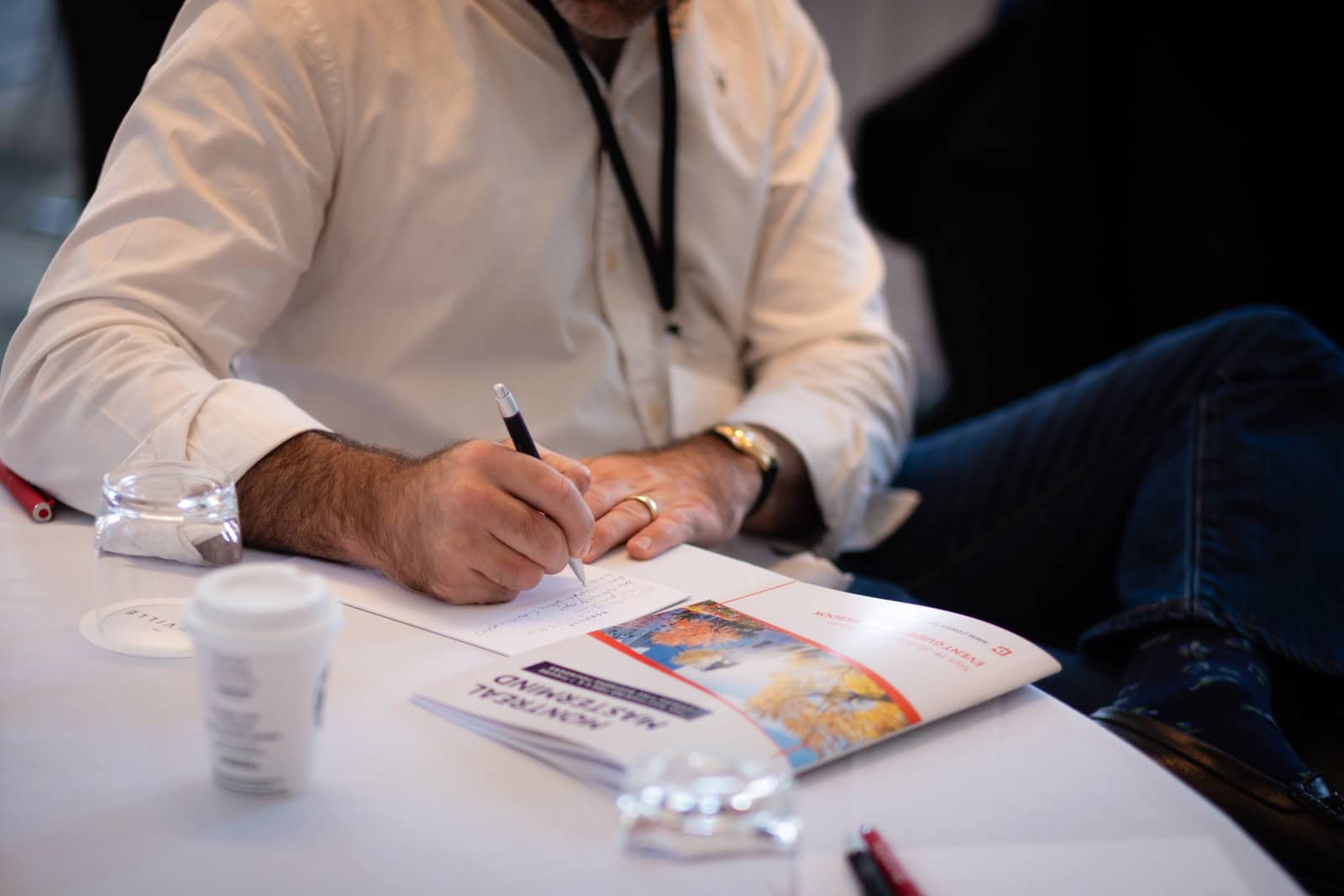 Consultant in a white shirt and event lanyard writing notes by hand at a Consulting Success Montreal Mastermind, with a coffee, a glass of water, and an event guide on the table.